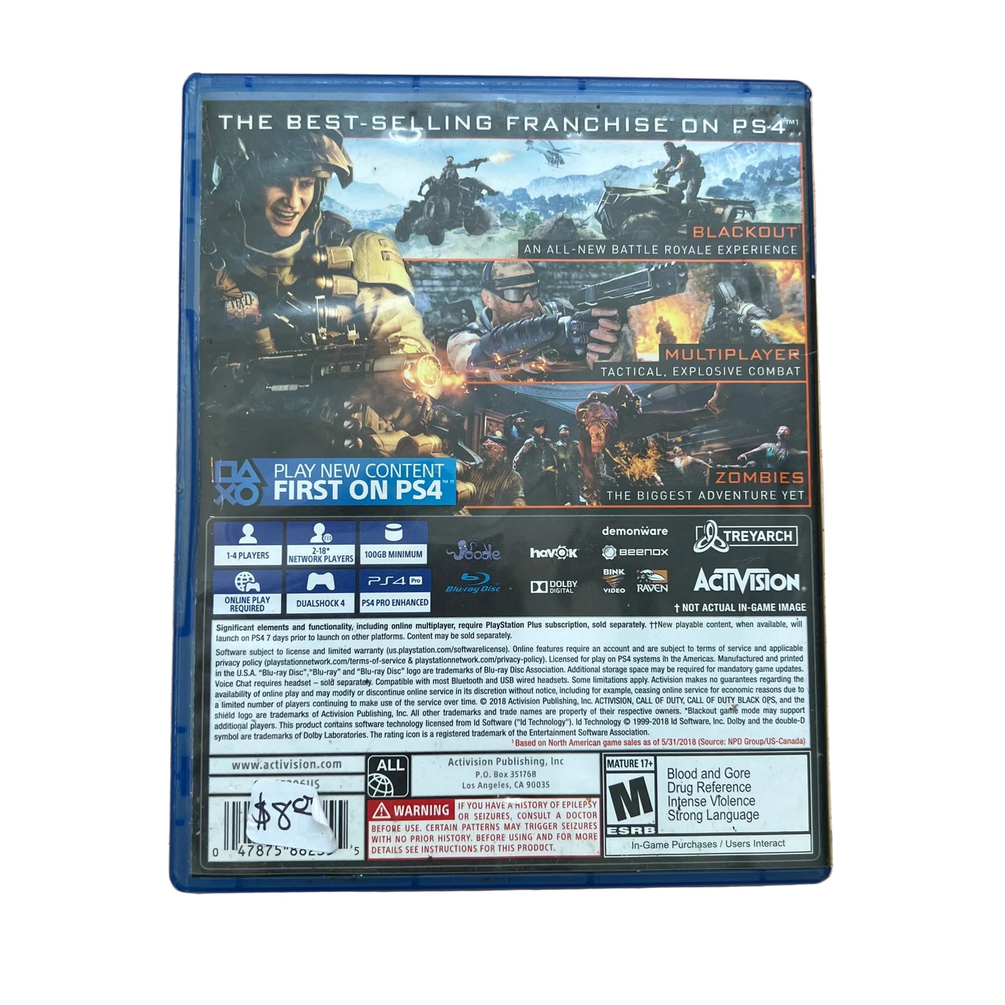 Back cover of the PS4 game 'Call of Duty: Zombie Warfare' resting on a light-colored wooden table. The blue plastic case shows the game's cover art, feature highlights, platform logos, and rating information.