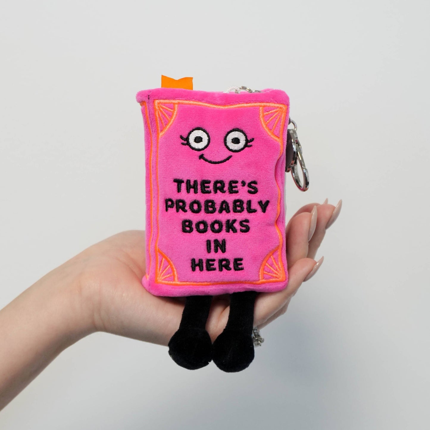 Pink plush book-shaped pouch with a face and text held by a hand against a light gray background